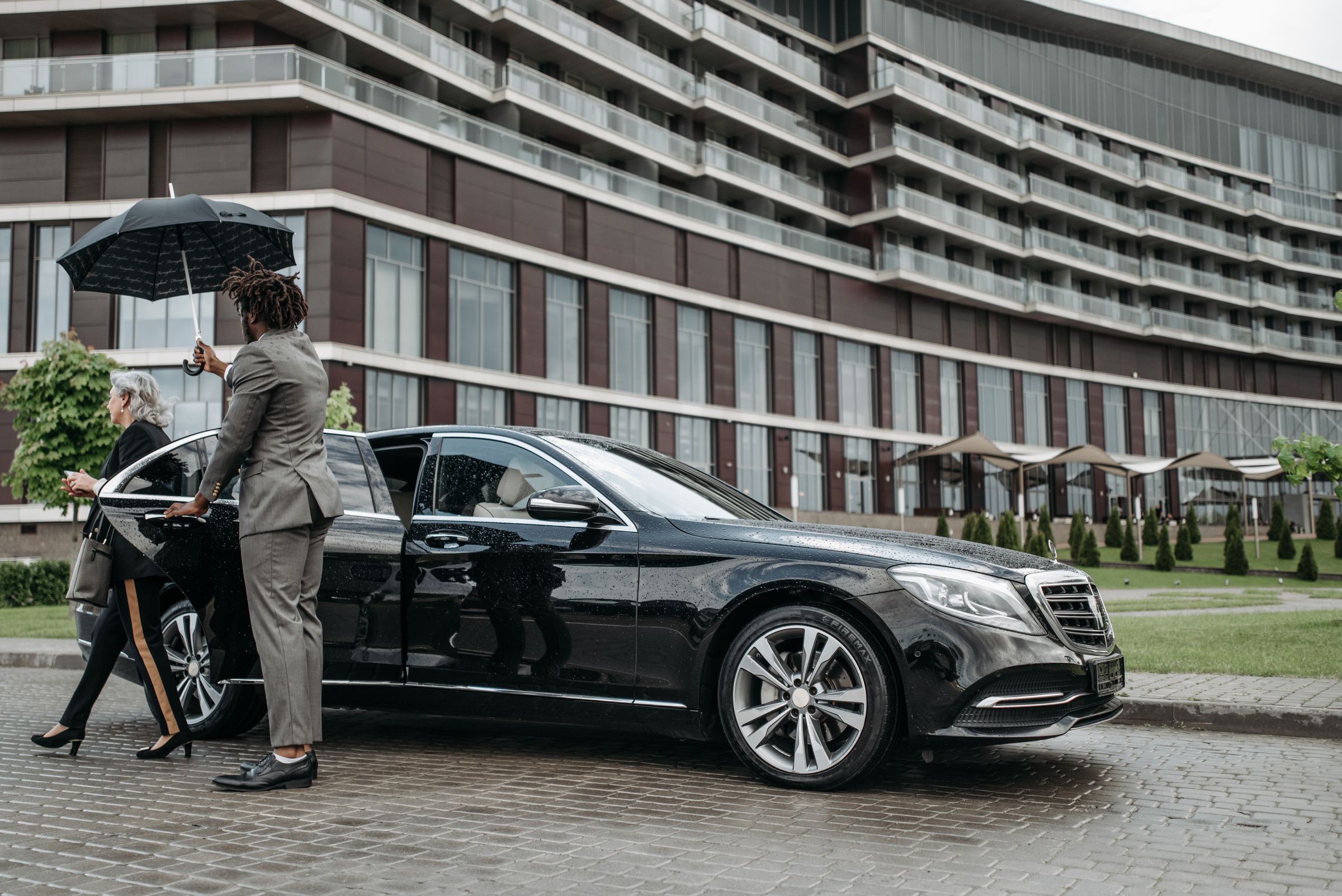 A suited close-protection professional assists an executive away from a black vehicle at a modern venue.