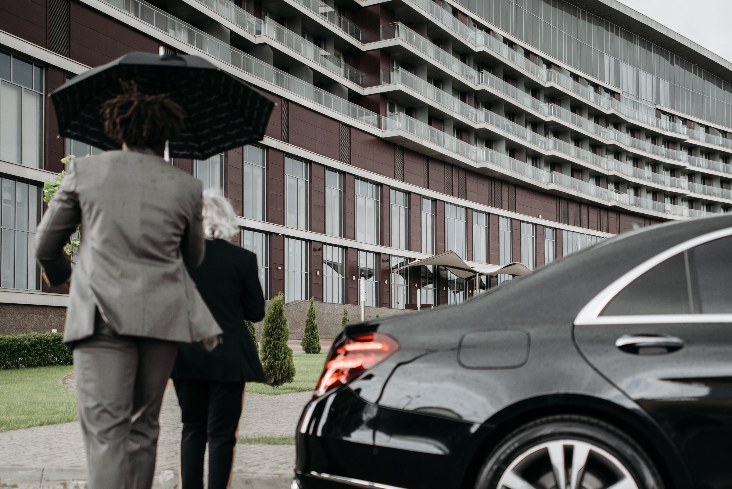 A suited protection professional escorts a principal from a black vehicle toward a modern building.